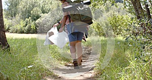 A person is hiking on a dirt trail carrying a large backpack and a white container, collecting trash