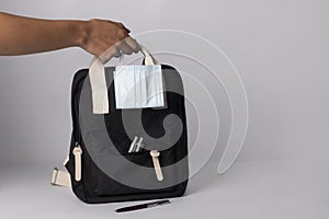 Person holding a black backpack with ballpens, and mask on white background - travel concept