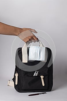 Person holding a black backpack with ballpens, and mask on white background - travel concept