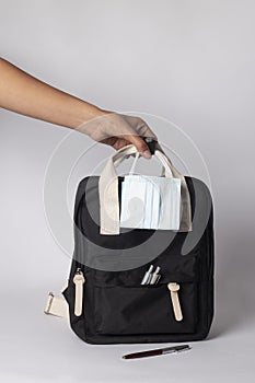 Person holding a black backpack with ballpens, and mask on white background - travel concept