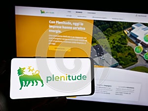 Person holding cellphone with logo of company Eni Plenitude SpA Societa Benefit on screen in front of business webpage.