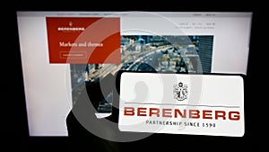 Person holding cellphone with logo of German bank Joh. Berenberg, Gossler Co. KG on screen in front of business webpage.