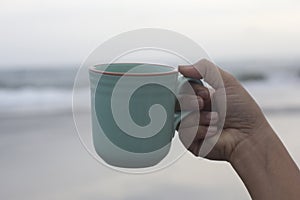 Person holding a cup of coffee in hand on beach background at sunset. Self love and care concept. Relax weekend