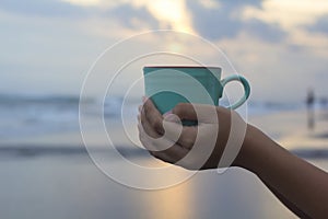 Person holding a cup of coffee in hand on beach background at sunset. Self love and care concept. Relax weekend