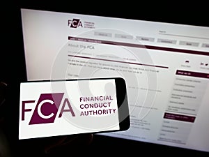Person holding mobile phone with logo of British Financial Conduct Authority (FCA) on screen in front of web page.