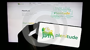 Person holding mobile phone with logo of company Eni Plenitude S.p.A. Societa Benefit on screen in front of web page.