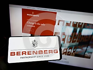 Person holding mobile phone with logo of German bank Joh. Berenberg, Gossler Co. KG on screen in front of web page.