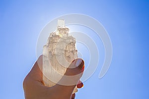 Person holding naturally carved mineral stone Selenite tower against sun and blue sky.