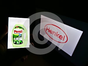 Person holding smartphone with laundry detergent product Persil on display marketed by company Henkel with logo in background.