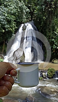 Person holding a traditional tea or coffee cup in hand against outdoor waterfall. Vertical composition