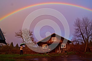 person holding an umbrella, rainbow arching over a quaint cottage