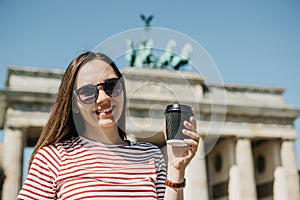 A person holds a disposable cup with coffee or another drink on the background of the Brandenburg Gate in Berlin.