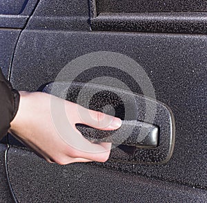 A person holds a frozen car handle in winter. A man opens the door in a black car covered in hoarfrost morning frosts.