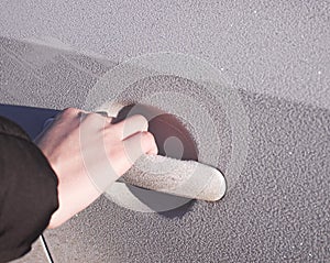 A person holds a frozen car handle in winter. A man opens the door in a white car covered in hoarfrost morning frosts.