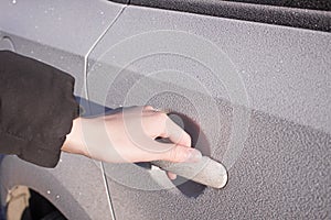 A person holds a frozen car handle in winter. A man opens the door in a white car covered in hoarfrost morning frosts.