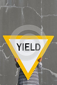 Person holds up big yield road sign. Bold yellow against cracked gray concrete wall.