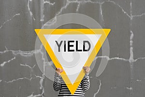 Person holds up big yield road sign. Bold yellow against cracked gray wall.