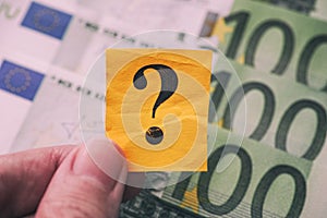 Person holds yellow paper note with question mark in his hand against Euro banknotes background