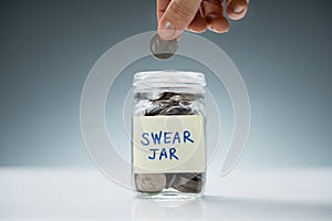 Person Inserting Coin In Swear Jar Over White Desk