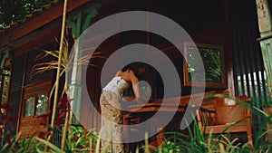 Person leaning on a wooden railing of a rustic house surrounded by lush greenery