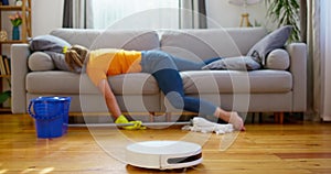 Person lying on sofa with mop, robotic vacuum cleaner on hardwood floor.