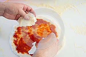 A person is making a pizza with tomato sauce and mozzarella cheese on a white worktop with flour sprinkled on it