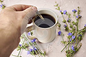 a person mixes a chicory drink with a spoon in a coffee mug next to chicory flowers