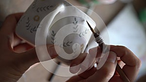 Person paints brown circles pattern on white ceramic mug in kitchen at home on blurred background