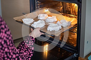 A person in a patterned shirt slides a tray of meringues into the oven, an intimate moment in home baking, perfect for