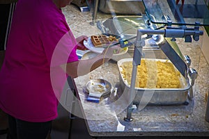 Person in pink teeshirt filling plate at breakfast buffet with scrambled eggs and waffle