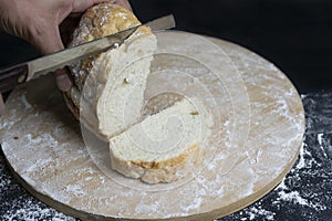 Person`s hand cutting wheat traditional bread with a knife on floury wooden board.Rustic bakery concept.Dark background