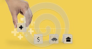 Person`s hand grabs a square wooden block showing an insurance icon placed against a wooden block, money icon, car and house.