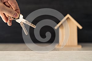 A person's hand holding the keys to their new home ownership with small wooden house in the background.