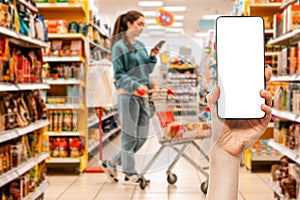 A person's hand holds a mobile phone. Mock up. In the background, a woman using cellphone is pushing a grocery cart
