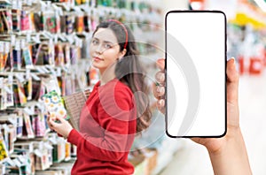 A person`s hand holds a mobile phone, with a shopping basket on the screen. In the background, a pretty woman selects digital