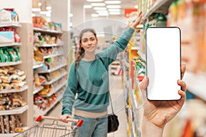 A person`s hand holds a mobile phone, with a shopping basket on the screen. In the background, a woman selects products in a