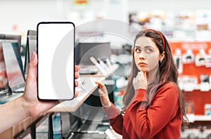 A person& x27;s hand holds a mobile phone, with a shopping basket on the screen. In the background, a woman thoughtfully