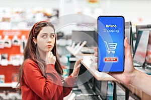 A person`s hand holds a mobile phone, with a shopping basket on the screen. In the background, a woman thoughtfully selects