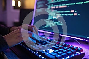 Person s hands typing on a glowing keyboard in front of a computer screen displaying code