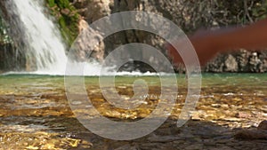 Person scoops drinking water in their hands from crystal clear river with waterfall, to quench thirst after a long walk