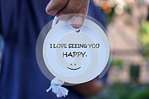 Person showing text on label paper in hand - I love seeing you happy. With smiling face emoticon. Kindness and happiness concept.