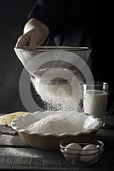 Person sifting flour into ceramic bowl. Dough preparation process. Milk, eggs, wooden table.