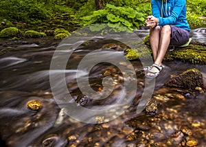 Person sits on the stone covered with moss in the center of rapid flow of the river, holding his feet in clear water