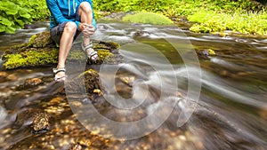 Person sits on the stone covered with moss in the center of rapid flow of the river, holding his feet in clear water