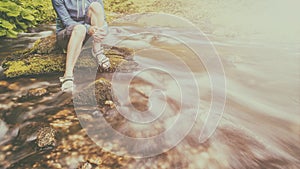 person sits on the stone covered with moss in the center of rapid flow of the river, holding his feet in clear water