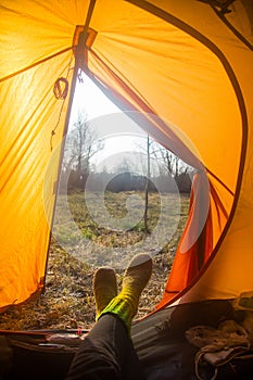 A person sitting in an orange tent, camp in the bank of the river in spring. Feet selfie of traveler. Relaxed, colorful look.