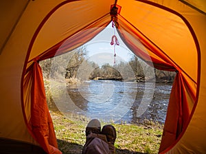 A person sitting in an orange tent, camp in the bank of the river in spring. Feet selfie of traveler. Relaxed, colorful look.