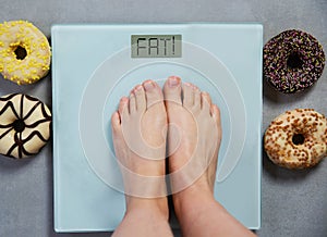 Person standing on digital weight scale showing the words FAT, and junk food donuts on background, diet concept