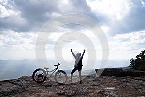 A person standing with hands up beside a bicycle on rocky mountain looking out at scenic