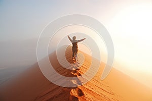 Person standing on top of dune in desert and looking at rising sun in mist with hands up, travel in Africa